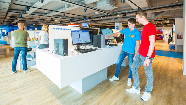 Un employé de Coolblue parle à un client d'un PC et d'un écran PC sur un meuble du magasin. Pendant ce temps, des clients regardent des produits en arrière-plan.