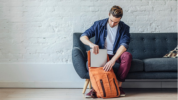 Un homme sur un canapé met un PC portable dans un sac à dos orange.