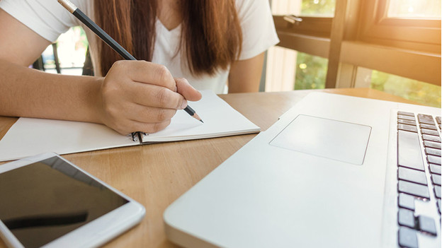 Woman writes in notebook while she works on a laptop behind a desk.