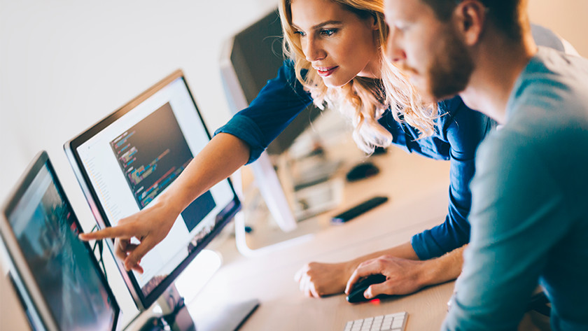 Woman points to monitor computer on desk. Man watches with computer mouse in hand.