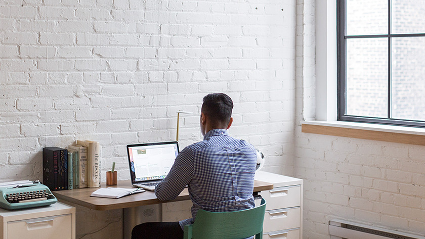 Man werkt achter bureau op zijn laptop met uitzicht op een muur. 