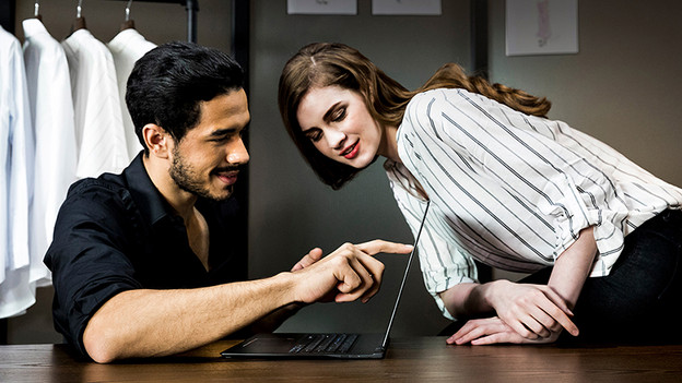 Man holds index finger on laptop screen and woman peeks around the corner.